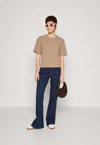 Braunfarbenes T-Shirt mit kurzen Ärmeln, dunkelblaue Schlagjeans, braune Handtasche und weiße Plateauschuhe; Model trägt weiße Sonnenbrille.