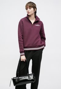 Tumši sarkans rāvējslēdzēja džemperis ar baltām svītrainām aprocēm, priekšpusē ar logotipu. Kombinēts ar melnu ādas somiņu, kas turēta rokā, un piegrieztām biksēm.