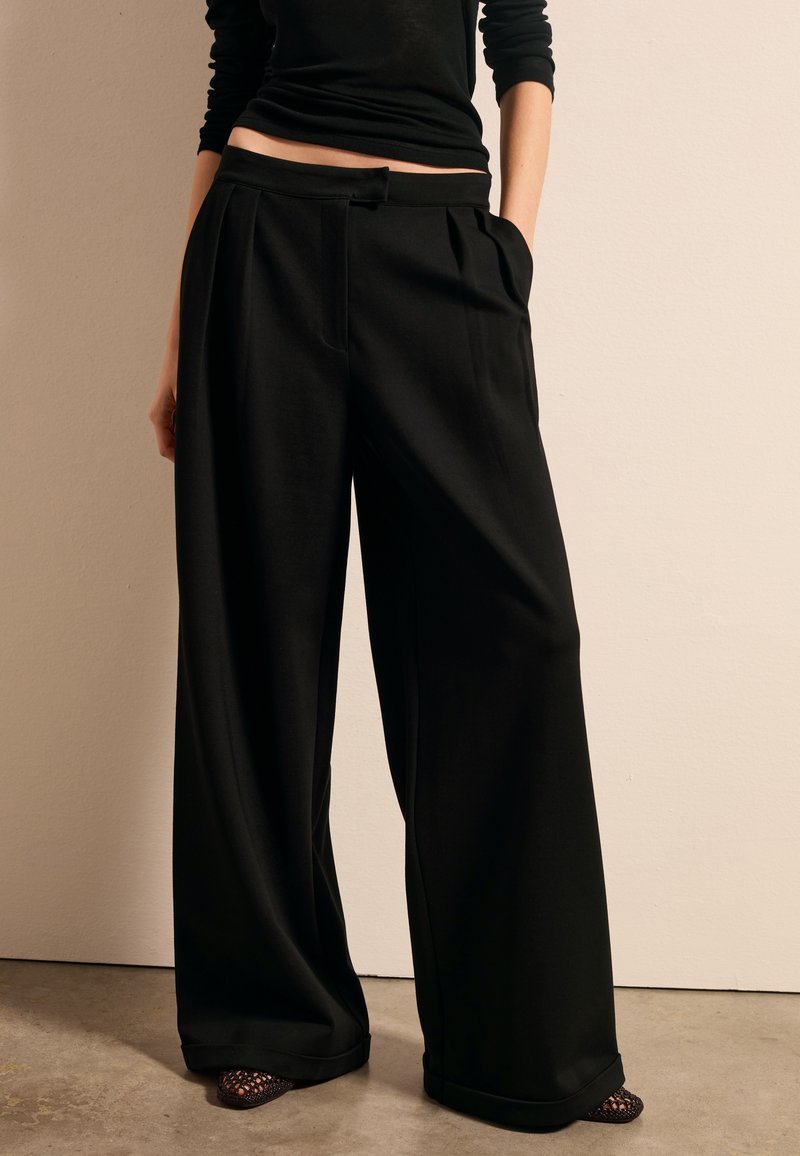 Pantalons noirs à jambe large avec des plis, fabriqués en tissu lisse. Dotés de poches et d'une taille cintrée. Portés avec des chaussures ouvertes.