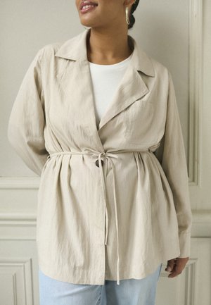 Chaqueta ligera beige con cuello, cintura ajustada, detalle de botones y mangas largas, llevada sobre una blusa blanca y combinada con vaqueros claros.