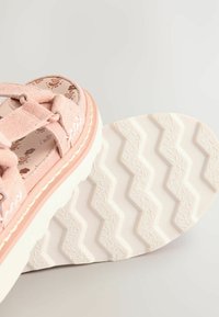 Sandale en daim rose avec des brides croisées, semelle extérieure en caoutchouc blanc avec un motif en zigzag, et design floral sur le coussin de pied.