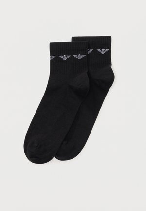 Paire de chaussettes noires pour cheville avec poignets côtelés et logo d'aigle blanc près du bord supérieur, présentées sur un fond clair uni.