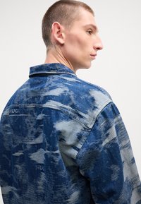 Blauwe spijkerjack met een vervaagd, textuurpatroon. Heeft een kraag en zichtbare stiksels. Bevat "HUGO" branding op de achterkant.