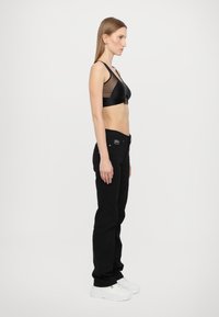 Crna mrežasta braletica s čvrstim remenom i crne traperice. Model nosi bijele tenisice. Pozadina je jednostavna bijela.
