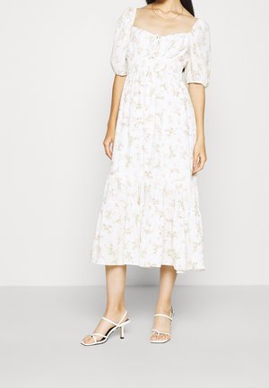 Femme portant une robe midi blanche à imprimé floral rose, manches courtes bouffantes, décolleté carré et sandales à talons blanches à lanières.