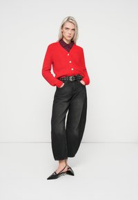 Punane kudumiskardigan, millel on V-kaelus ja viis valget nööpi, koos laia musta teksapüksiga ja mustade terava ninaga kingadega.