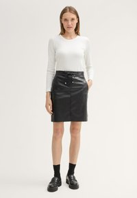 Schwarzer Lederrock mit Kordelzugbund und seitlichen Taschen, kombiniert mit einem weißen Langarmshirt und schwarzen Schuhen mit klobigen Sohlen.