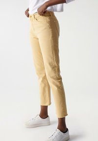 Jeans cortos de color amarillo claro, con un ajuste de pierna recta, que presentan un dobladillo sin terminar y bolsillos frontales, combinados con zapatillas blancas.