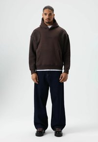 Bruine hoodie met gestructureerde accenten en een voorzak, gecombineerd met blauwe wijde jeans en zwarte veterschoenen. Simpel ontwerp, minimale branding.