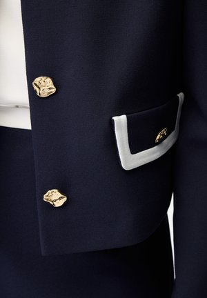 Marineblauwe stof met twee onregelmatige gouden knopen en een marineblauw zakklepje afgezet met wit en een kleine gouden decoratieve knoop.