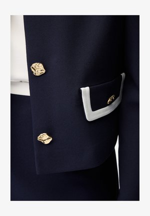 Marineblauwe stof met twee onregelmatige gouden knopen en een marineblauw zakklepje afgezet met wit en een kleine gouden decoratieve knoop.