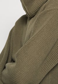 Pull à capuche tricoté vert olive avec tissu texturé, doté d'un col montant et d'une coupe décontractée. Détails côtelés visibles le long des coutures.