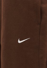 Καφέ ύφασμα με λευκό κεντημένο λογότυπο Nike swoosh κοντά σε κάθετη ραφή, πιθανώς μέρος ενδύματος.