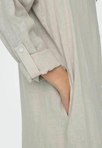 Camicia in lino beige leggera con maniche arrotolate, dettagli con bottoni e tasche laterali. Il tessuto presenta un motivo testurizzato sottile.