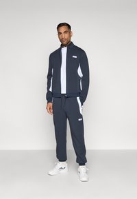 Navy treningsdrakt i mykt stoff med hvite sidepaneler, jakke med glidelås og høy krage, samt bukser med mansjetter. Avsluttet med logo-detaljer.