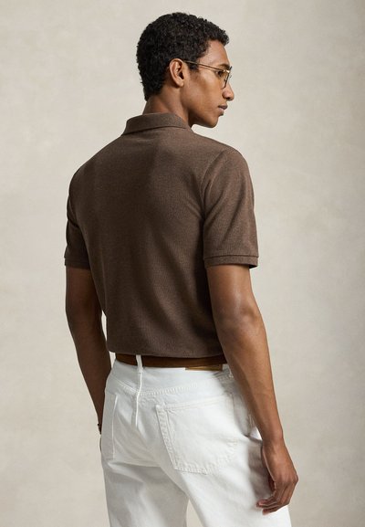 Mees, kes kannab pruuni lühikeste varrukatega polo särki ja valgeid pükse, pööratud küljele, pea pööratud paremale ühtlase tausta ees.