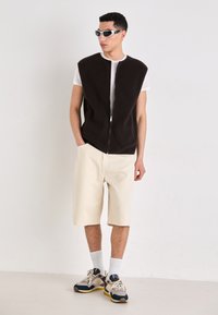 Chaleco de forro polar negro con cremallera en la parte delantera, llevado sobre una camiseta blanca. Combinado con pantalones cortos color crema y zapatillas deportivas. Los accesorios incluyen calcetines blancos y gafas de sol.