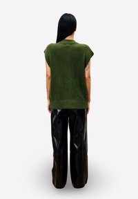 Vrouw met lang zwart haar, gekleed in een groene mouwloze gebreide top en zwarte wijde leren broek, met haar rug naar de toeschouwer, op een witte achtergrond.