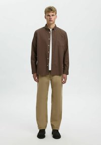 Camisa marrón de botones con cuello, llevada sobre una camiseta blanca. Pantalones de pierna ancha beige y zapatos negros completan el conjunto.