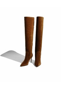 Botas de ante marrón hasta la rodilla con puntas afiladas y altos tacones de aguja. Textura suave con un diseño ajustado. Sombra visible en el suelo.