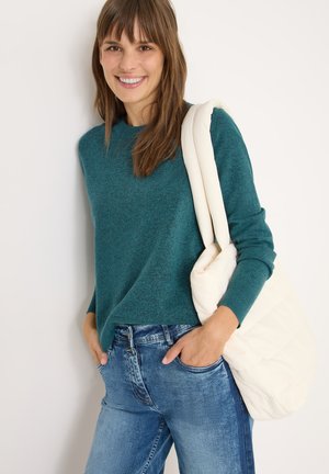 Maglione turchese con un finish testurizzato, abbinato a jeans azzurri chiari. Porta una borsa cremosa, morbida e trapuntata. Design semplice e vestibilità rilassata.