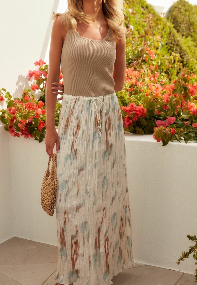 Femme portant un débardeur beige et une longue jupe plissée avec des motifs bleus et marron, tenant un sac tressé, debout près de plantes en fleurs.