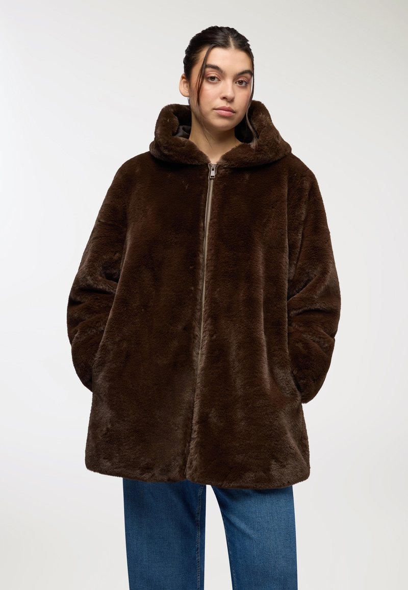 Giacca con cappuccio in pelliccia sintetica marrone, con chiusura a zip, design oversize e tasche laterali. Texture morbida e silhouette rilassata, indossata sopra un denim.