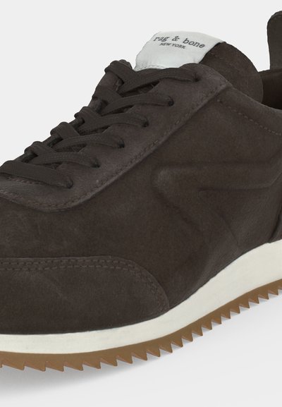 Gros plan sur une basket en daim marron foncé avec des lacets assortis, une semelle intermédiaire blanche, une semelle extérieure marron crantée et une étiquette "rag & bone New York" sur la languette.
