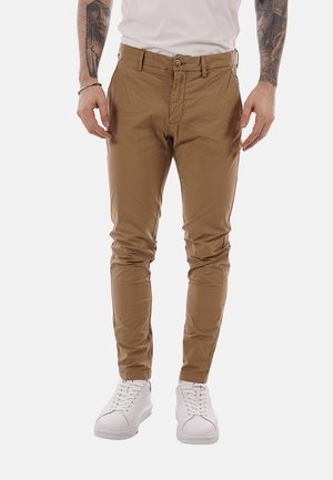 Uomo che indossa pantaloni marroni slim fit, sneakers bianche e una camicia bianca, visibile dalla metà del busto ai piedi con gli avambracci tatuati in vista.