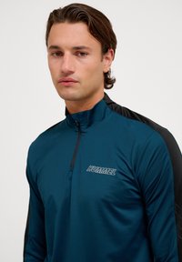 Teal långärmad atletisk topp med svart accent på axeln, har en hög krage och en fjärdedels dragkedja. "HUMMEL"-logotypen tryckt i vitt.