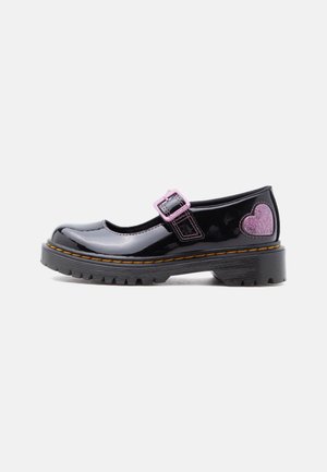 Chaussure Mary Jane noire en cuir verni avec boucle violette et cœur violet scintillant sur le talon, semelle épaisse avec surpiqûres jaunes.