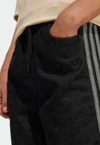 Pantaloni jogger in denim nero con vita con coulisse, tre strisce verticali grigie e un piccolo logo nero vicino alla tasca. La texture appare leggermente spazzolata.