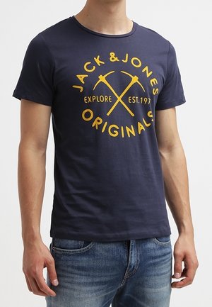 Mand iført en navyblå t-shirt med gule krydsede hakke-værktøjer og tekst "Jack & Jones Originals Explore Est. 1975" foran.