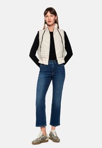 Gepolsterte Weste in hellem Creme mit einem strukturierten Wellendesign, kombiniert mit einem schwarzen Oberteil und blauen Jeans. Dazu Zebra-Muster-Sneaker.