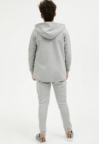 Grau Hoodie und passende Jogginghose aus weichem Material; verfügt über eine Kapuze, lange Ärmel und enge Bündchen. Die Schuhe sind graue Sneakers mit orangefarbenen Akzenten.