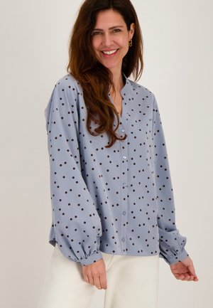 Vrouw glimlachend, gekleed in een lichtblauwe blouse met lange mouwen, zwarte stippen en een witte broek, voor een effen achtergrond.