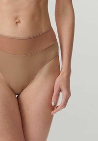 Primo piano di una persona che indossa biancheria intima seamless beige a vita alta, mostrando il torace inferiore, i fianchi e il braccio destro su uno sfondo neutro.