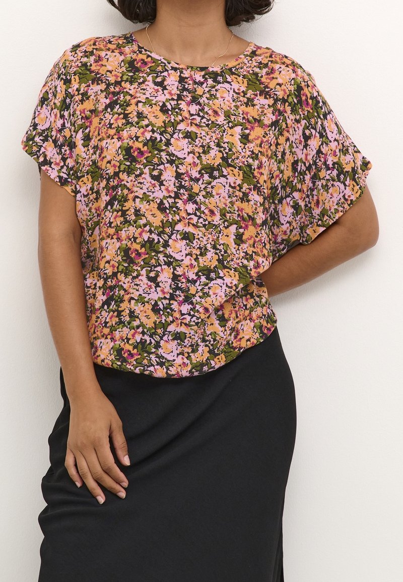 Blouse fleurie aux motifs vibrants rose, orange et vert. Manches courtes, coupe ample, encolure arrondie, portée sur une jupe noire.