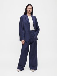 Femme debout, main dans la poche, portant un blazer bleu marine ample, un pantalon large bleu marine, un haut blanc et des chaussures pointues de couleur claire sur fond blanc.