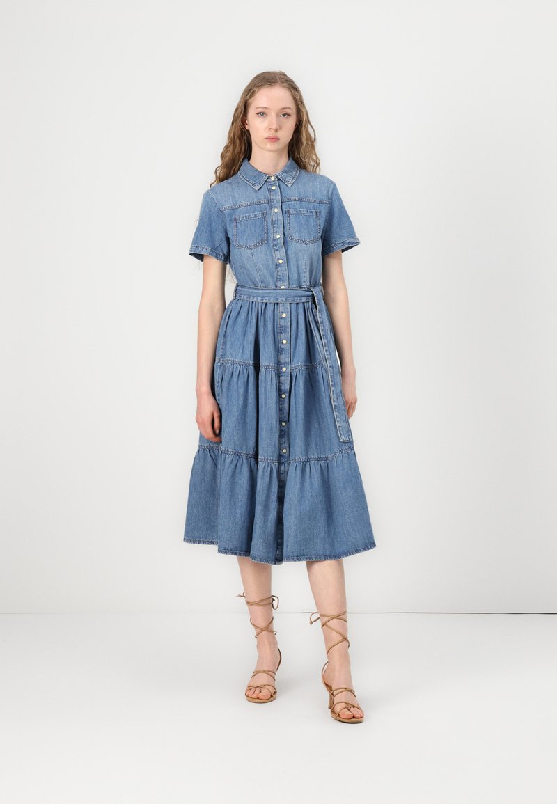 Robe en denim à boutons avec manches courtes, deux poches poitrine, taille ceinturée, jupe à volants et ourlet au genou, de couleur bleu clair.