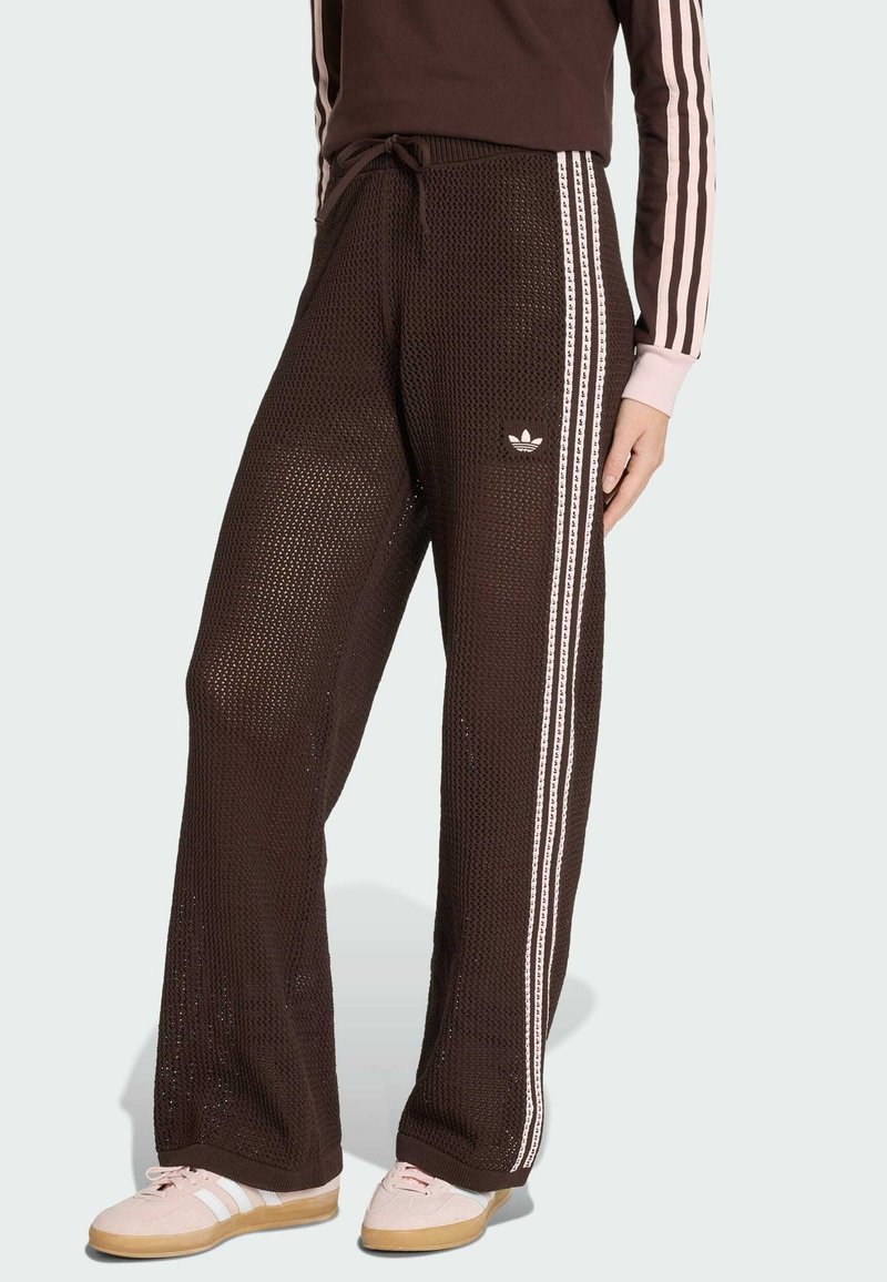 Henkilö pukeutuneena ruskeisiin neulostettuihin Adidas-housuihin, joissa on valkoiset kolme raitaa sivuilla, ja vastaavaan ruskeaan yläosaan, jossa on vaaleanpunaiset raidat hihoissa.