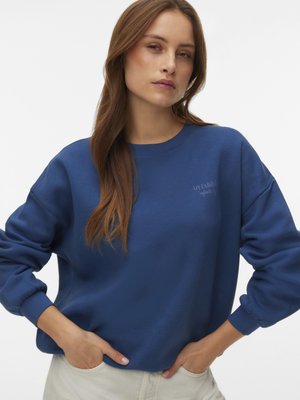Femme aux longs cheveux bruns portant un sweat-shirt bleu avec un texte discret sur le côté gauche de la poitrine, mains dans les poches avant, sur un fond blanc uni.
