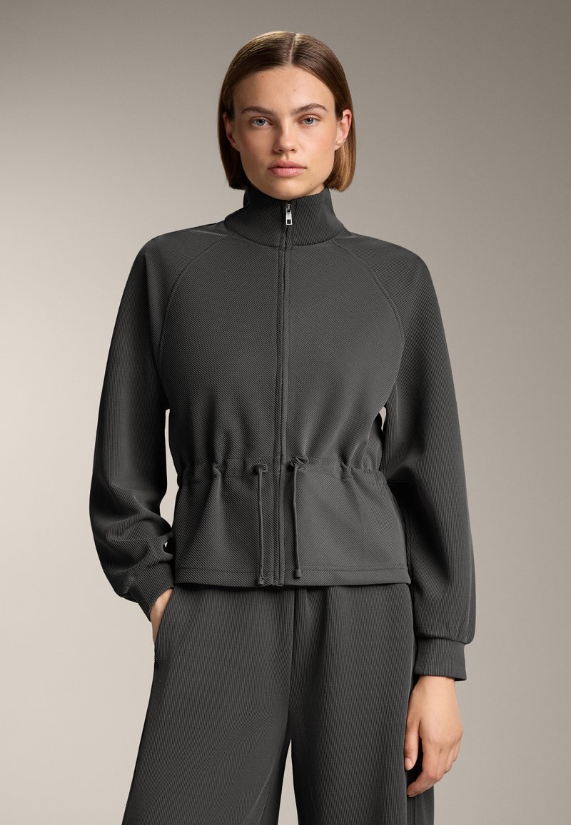 Jachetă cu fermoar de culoare gri închis, cu guler înalt, talie elastică și textură ribbed. Dispune de mâneci lungi și un croi lejer.