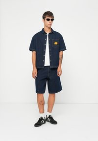 Camisa de botón de manga corta de mezclilla azul oscuro con bolsillo en el pecho y logo, combinada con pantalones cortos de mezclilla a juego y zapatillas deportivas negras.