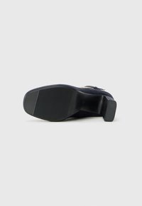 Chaussure à talon pour femmes, semelle noire avec détail de boucle, présentée vue de la semelle sur un fond gris clair uni.