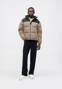 Jachetă căptușită maro și neagră, cu guler înalt, cu fermoar în față; asortată cu pantaloni din corduroy închiși la culoare și adidași albi. Textură netedă.