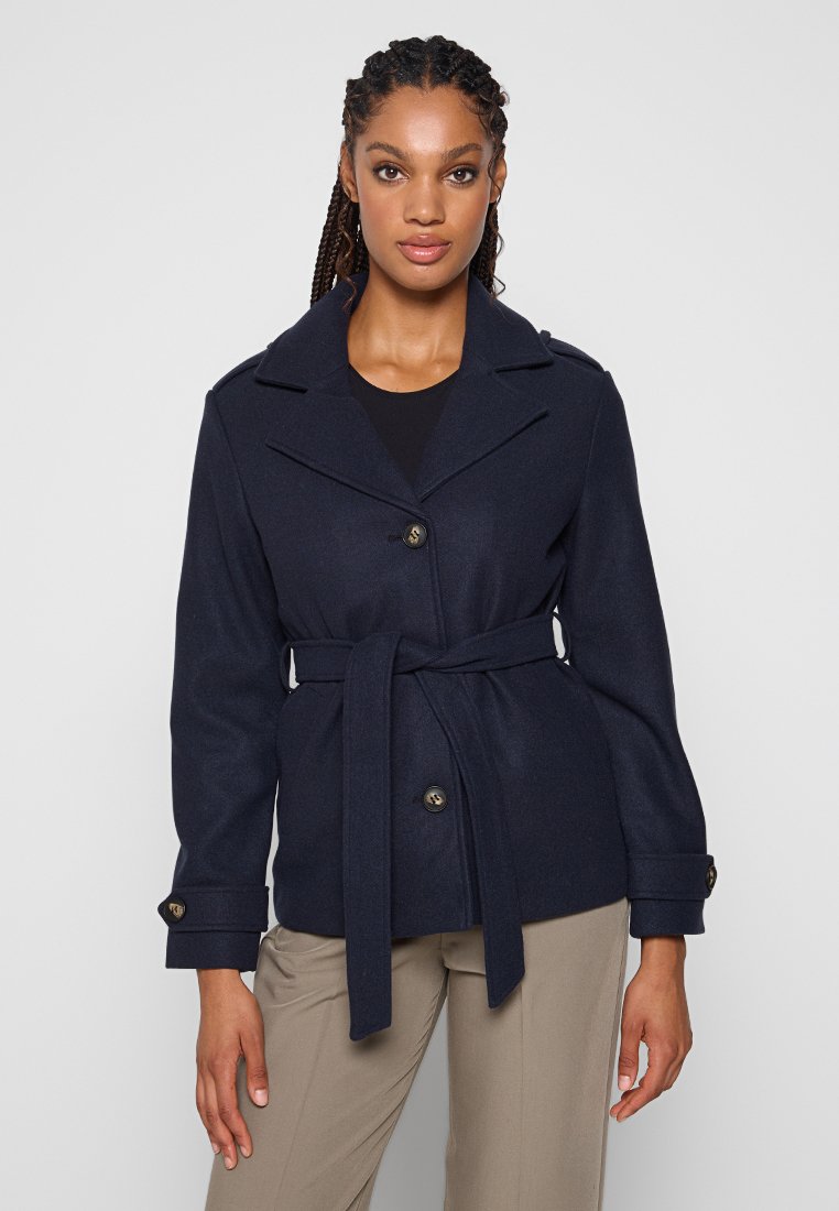 Femme portant un manteau ceinturé bleu marine avec des boutons, un haut noir et un pantalon beige, se tenant devant un fond clair uniforme.
