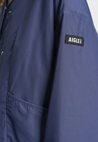 Veste bleu marine avec fermeture à boutons et poche latérale, présentant un patch noir sur la manche avec le texte blanc "AIGLE 1853".