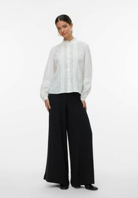 Witte blouse met gestructureerde stippen, gerimpelde kraag en verticale plooidetails, gecombineerd met brede zwarte broeken en zwarte schoenen.