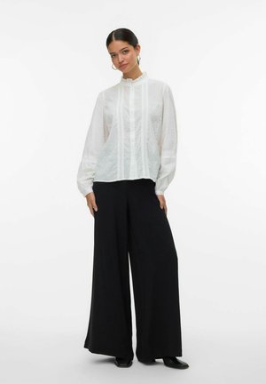 Witte blouse met gestructureerde stippen, gerimpelde kraag en verticale plooidetails, gecombineerd met brede zwarte broeken en zwarte schoenen.